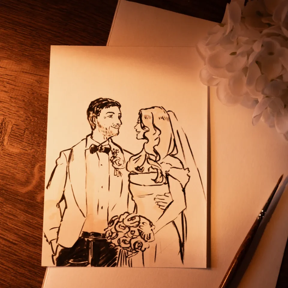 Dessin à l'encre noire d'un couple de mariés sur papier blanc, entouré d'un pinceau et de fleurs.